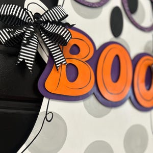 Puede incluir: Decoraci&oacute;n de fantasma de madera con tem&aacute;tica de Halloween, con la palabra "BOO" en letras naranjas con contornos negros. El fantasma tiene gafas moradas con purpurina y un lazo a rayas blanco y negro. El cuerpo del fantasma es blanco con lunares grises.