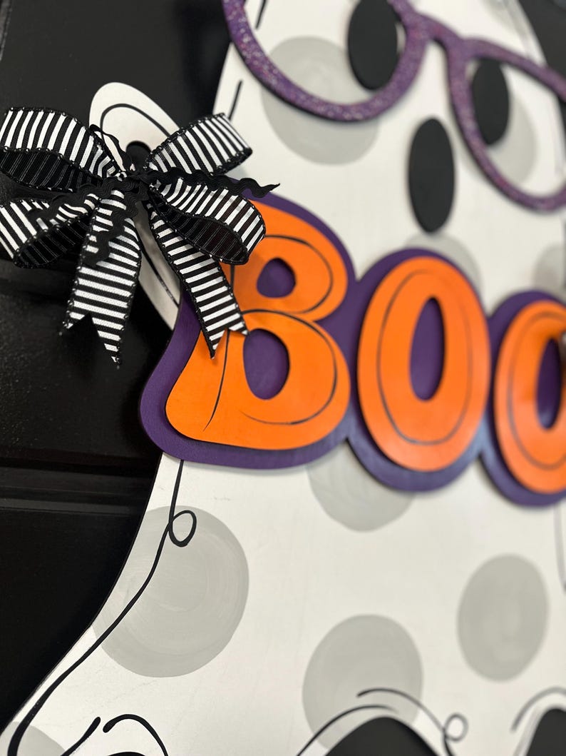 Puede incluir: Decoraci&oacute;n de fantasma de madera con tem&aacute;tica de Halloween, con la palabra "BOO" en letras naranjas con contornos negros. El fantasma tiene gafas moradas con purpurina y un lazo a rayas blanco y negro. El cuerpo del fantasma es blanco con lunares grises.