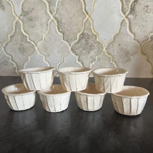 May include: Six white ceramic ramekins with a textured, leaf-like design. The ramekins are arranged in a staggered pattern on a black surface.