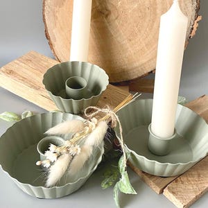 Könnte beinhalten: Salbeigrüne Kerzenhalter mit geriffeltem Design, die hohe weiße Kerzen halten. Die Halter sind auf Holzblöcken angeordnet, mit Trockenblumen und Grünzeug. Eine rustikale, minimalistische Ästhetik. Kerzen ca. 20 cm hoch.