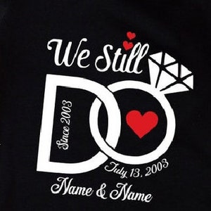 Könnte beinhalten: Schwarzes T-Shirt mit einem weißen Design aus zwei ineinandergreifenden Ringen mit einem Diamantring oben. Der Text "We Still Do" befindet sich über den Ringen. Der Text "Since 2003" befindet sich in einem Ring und "July 13, 2003" im anderen Ring. Der Text "Name & Name" befindet sich unter den Ringen.