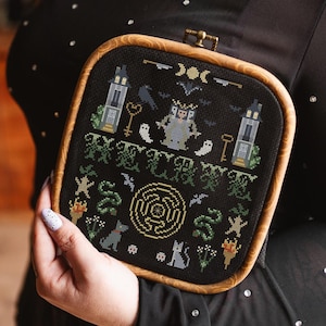 May include: A framed cross-stitch featuring a Halloween theme. The black fabric displays a design with a castle, keys, ghosts, bats, and a maze. The wooden frame is square with rounded corners.