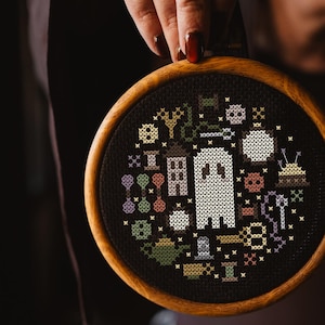 May include: A cross-stitch embroidery in a wooden hoop, featuring a ghost and Halloween motifs. The design is stitched on black fabric. The colour palette includes white, green, purple, and yellow. The hoop is approximately 15 cm in diameter.