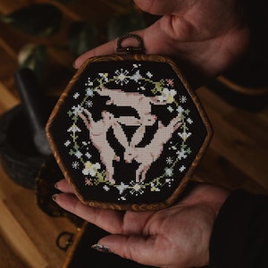 May include: A hexagonal wooden frame with a black fabric background featuring an embroidered design of three white rabbits hopping in a circle surrounded by white flowers and stars.