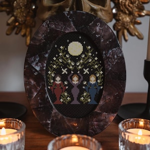 May include: A framed cross-stitch embroidery depicting three figures standing under a tree with a full moon in the background. The frame is oval-shaped and made of dark brown wood with a mottled finish. The figures are wearing long robes in shades of red, purple, and blue. The tree is green with white flowers and the moon is yellow.
