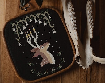 May include: A framed cross-stitch embroidery depicting a deer with antlers standing under a canopy of white flowers and candles. The embroidery is on black fabric and is framed in a dark wood frame.