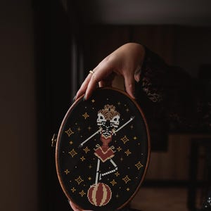 May include: A black embroidery hoop with a cross-stitch design of a skeleton in a red and gold circus costume balancing on a ball. The background is black with gold stars.