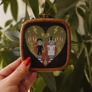 Puede incluir: Una bordado de punto de cruz enmarcado que presenta un diseño de corazón con dos figuras en su interior. El corazón es verde y amarillo con un fondo negro. Las figuras son estilizadas y visten ropa negra y blanca. El marco es de madera marrón.