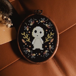 May include: Oval cross-stitch embroidery in a wooden hoop. The design features a light gray figure with a black background, surrounded by leaves and insects. The embroidery is set against a brown leather surface.