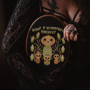 Puede incluir: Un bastidor de bordado de punto de cruz negro con marco de madera. El diseño presenta una figura con cabeza de calabaza, tres figuras más pequeñas y mazorcas de maíz, con el texto "WHAT A WONDERFUL HARVEST". La paleta de colores incluye naranja, verde y marrón.