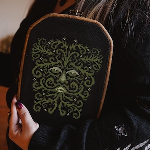 May include: A black fabric pouch with a wooden frame, featuring a green embroidered design of a face with a beard. The design is reminiscent of a forest spirit or nature deity.