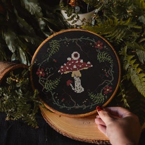Puede incluir: Un bastidor de bordado circular de madera que contiene un diseño de punto de cruz sobre tela negra. El diseño presenta un hongo central rojo y blanco con un solo ojo en el sombrero, rodeado por una corona de helechos verdes, pequeñas flores rojas y estrellas.