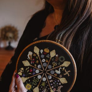 Può includere: Un cerchio da ricamo nero con un disegno a punto croce colorato che presenta una varietà di simboli di strega, tra cui scope, gufi, lune e stelle. Il disegno è su uno sfondo nero ed è circondato da un cerchio di legno.