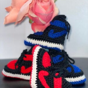 May include: Crocheted baby sneakers in black, red, and blue. The sneakers have white soles and laces, with a blue Nike swoosh on one pair and a red swoosh on the other. A pink rose is in the background.