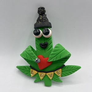 Könnte beinhalten: Grünes Marihuana-Blatt mit einer schwarzen Mütze, Kulleraugen, einem roten Herzen und einem goldenen Banner mit der Aufschrift "LOVE".