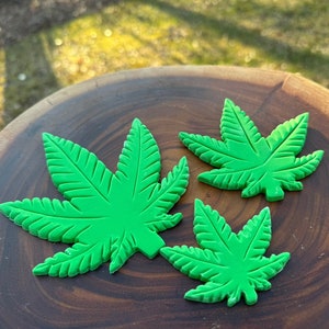 Könnte beinhalten: Drei grüne Keksstempel in Form von Marihuana-Blättern auf einer Holzoberfläche.