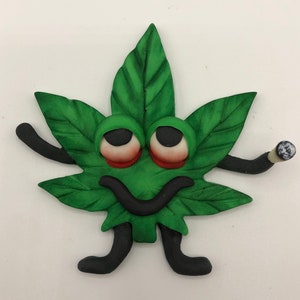 Könnte beinhalten: Ein grünes Marihuana-Blatt mit einem lächelnden Gesicht, Kulleraugen und schwarzen Armen und Beinen. Das Blatt hält in seiner rechten Hand einen kleinen, gerollten Joint.