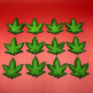 Könnte beinhalten: Zwölf grüne Cannabisblatt-Dekorationen, angeordnet in einem Raster auf rotem Hintergrund. Die Blätter haben eine strukturierte Oberfläche mit sichtbaren Adern und einer dunkleren grünen Schattierung. Die Anordnung ist symmetrisch.