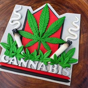 Könnte beinhalten: Ein dekoratives Cannabis-Design mit einem großen grünen Cannabisblatt auf rotem Hintergrund, zwei angezündeten Joints und dem Wort "CANNABIS" in silbernen Buchstaben. Das Design umfasst grüne Cannabisblätter und weiße Rauchfahnen.