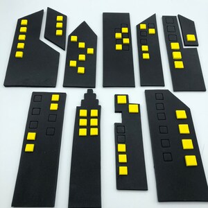 Pode incluir: Um conjunto de dez recortes de edif&iacute;cios da cidade de feltro preto com janelas amarelas. Os edif&iacute;cios s&atilde;o de v&aacute;rios tamanhos e formas.