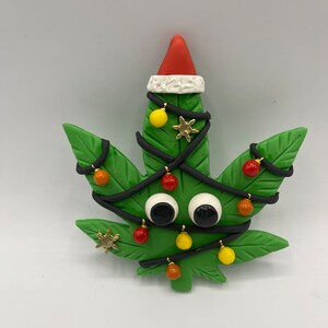 Könnte beinhalten: Ein grüner Weihnachtsbaum-Anhänger in Form eines Marihuana-Blattes mit einer roten Weihnachtsmütze, schwarzen Lichterketten, roten und gelben Kugeln und Glitzern.