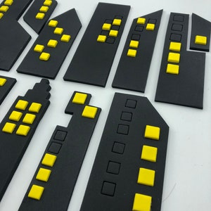 Pode incluir: Silhuetas de cidade de espuma preta com janelas amarelas. As silhuetas est&atilde;o dispostas em fila, com o edif&iacute;cio mais alto no centro.