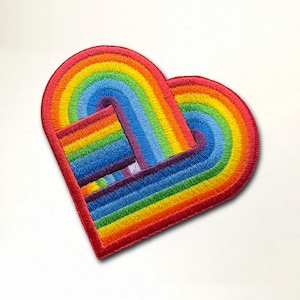 Op de afbeelding: Een regenboogvormige hartvormige patch met een gelaagd regenboogontwerp. De patch is gemaakt van vilt en heeft een rode rand.