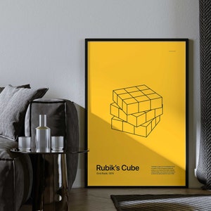 May include: A black framed poster with a yellow background and a black line drawing of a Rubik's Cube. The text "Rubik's Cube" and "Ernő Rubik, 1974" is printed in black at the bottom of the poster.