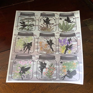 May include: Nine glass jars with lids, each containing a different type of dried flowers and a silhouette of a fairy. The jars are arranged in three rows of three, and the background is a light blue with clouds.