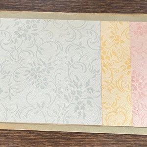 May include: Three samples of wallpaper with a floral pattern. The first sample is a light gray with a silver floral pattern. The second sample is a light yellow with a gold floral pattern. The third sample is a light pink with a pink floral pattern.
