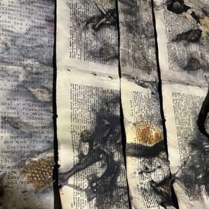 May include: A close-up of a book with black ink splattered across the pages. The book is folded in half and the pages are slightly curled. The text on the pages is mostly obscured by the ink, but some words are still visible. The book appears to be old and worn.