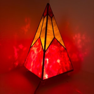 Könnte beinhalten: Eine Pyramide aus Buntglas mit roten, gelben und orangen Glasscheiben. Die Lampe ist eingeschaltet und strahlt ein warmes Licht aus.