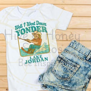 Puede incluir: Camiseta blanca con un gráfico de un hombre esquiando en el agua y el texto "Well I Went Down Yonder to Lake Jordan". Se muestra con unos shorts vaqueros desgastados. El conjunto está sobre una superficie de madera.
