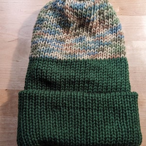 May include: A knitted beanie hat with a folded brim. The hat features a color block design with a dark green brim, a light tan stripe, and a top section with blue, green, and brown variegated yarn.