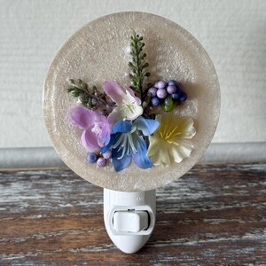 May include: A decorative night light featuring a round, beige resin base adorned with colorful artificial flowers and greenery. The flowers include purple, blue, and yellow blooms. The night light has an on/off switch and a standard plug.