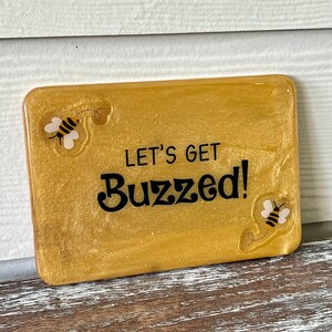 May include: A gold glitter resin sign with the text "Let's Get Buzzed!" and two cartoon bees on either side.