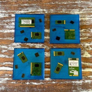 May include: Four square blue coasters with embedded computer components. Each coaster features a different arrangement of green circuit boards and black chips. The coasters are set against a wooden background.