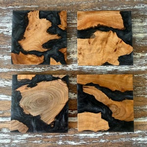 May include: Four square wooden coasters with a unique design. Each coaster features a natural wood grain pattern with a dark, black resin filling the spaces between the wood pieces. The coasters are arranged on a distressed, light-colored wooden surface.