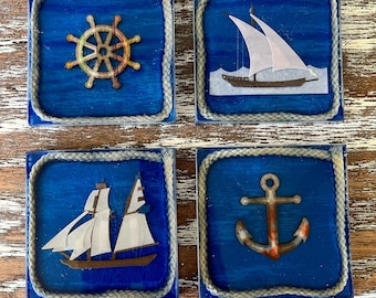 May include: Set of four nautical-themed coasters. Each square coaster features a blue background with a different design: a ship's wheel, a sailboat, a two-masted ship, and an anchor. The coasters are framed with a rope-like border.