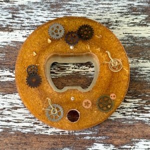 May include: A round, amber-colored bottle opener with embedded gears and gold flakes. The opener has a central cutout for leverage and is decorated with various sizes and styles of metallic gears.