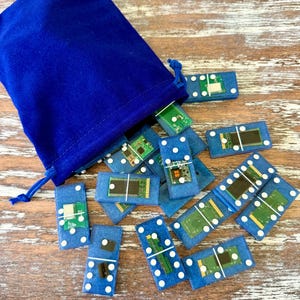 May include: A collection of blue dominoes with white dots and embedded circuit board designs, spilling from a matching blue velvet drawstring bag. The dominoes are scattered on a wooden surface.