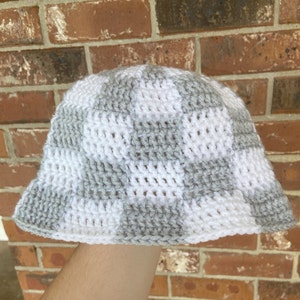 May include: Handmade crochet bucket hat in a checkerboard pattern of white and gray. The hat is made of soft yarn and has a slightly flared brim. A stylish accessory for any casual outfit.
