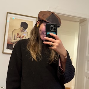 May include: A brown and black leather newsboy cap. The cap has a rounded crown and a short, stiff brim. The person in the image is wearing a black sweater and gold earrings. The person is holding a black phone in front of their face.