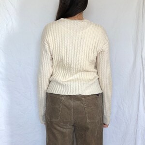 May include: A white cable knit sweater with long sleeves and a v-neck. The sweater is paired with brown corduroy trousers.