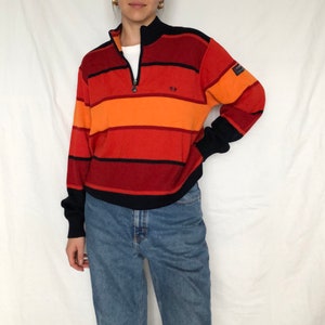 Può includere: Un maglione a righe con una chiusura a zip a metà. Il maglione ha righe orizzontali in tonalità di rosso, arancione e nero. Il maglione ha un piccolo logo sul davanti.