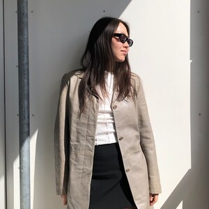 Puede incluir: Una mujer con una chaqueta larga de manga larga de color gris claro, una minifalda negra y gafas de sol negras.