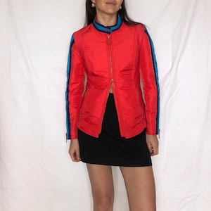 Könnte beinhalten: Eine rote Reißverschlussjacke mit blauen und weißen Streifen an den Ärmeln. Die Jacke hat einen Stehkragen und eine figurbetonte Silhouette.