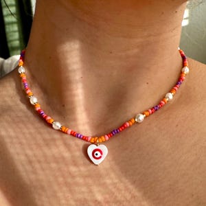 May include: A colorful beaded necklace with a heart pendant. The necklace features a mix of orange, pink, and purple beads, interspersed with white pearls. The pendant is a white heart with a red and black eye design.