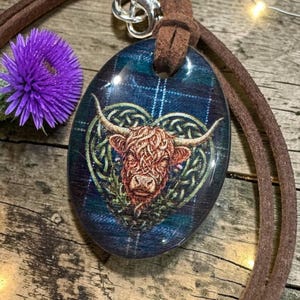 May include: An oval pendant featuring a Highland cow illustration centred within a green Celtic knot heart design, set against a blue and green plaid background. The pendant hangs from a brown suede cord with a silver-toned metal clasp.