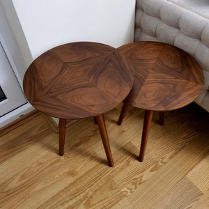 May include: Two round side tables with a dark brown, geometric patterned tabletop. Each table has three slender, tapered legs. The tables are placed on a light-coloured wooden floor.